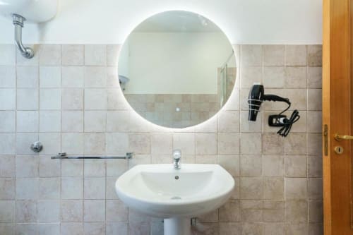 Bagno moderno nell'appartamento Origenes Rooms, Procida, Italia con specchio rotondo illuminato, lavabo a colonna e piastrelle beige