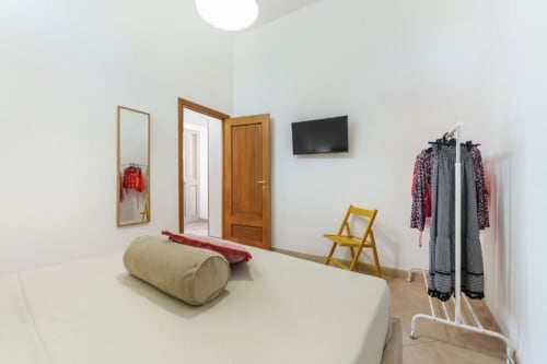 Camera moderna nell'appartamento di Procida con letto beige, porte in legno, sedia gialla, TV, specchio, attaccapanni e pavimenti chiari