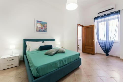 Camera luminosa con letto turchese, comodini bianchi, tende blu e pavimento in piastrelle presso Origenes Rooms a Procida