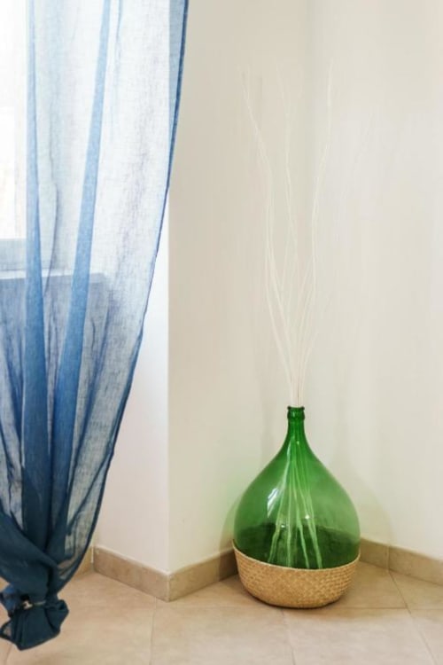 Vaso in vetro verde con erbe secche in cesto intrecciato sul pavimento piastrellato vicino a tenda blu, Origenes Rooms, Procida