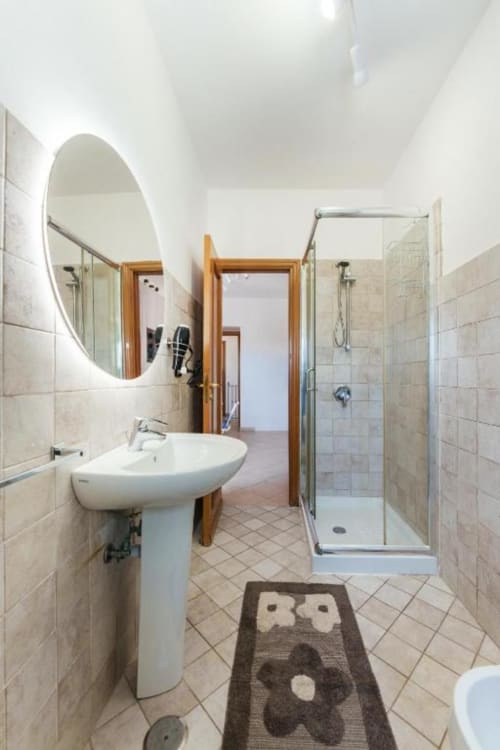 Bagno moderno nell'appartamento di Procida con specchio circolare illuminato, lavabo su piedistallo, doccia in vetro e pavimento in cotto
