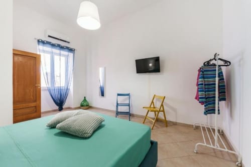 Camera luminosa degli Origenes Rooms a Procida con letto verde menta, porta in legno, tende blu, TV, sedie e appendiabiti