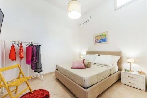 Camera luminosa dell'appartamento di Procida con letto beige, sedia gialla, appendiabiti colorato e opere d'arte con vista sul mare