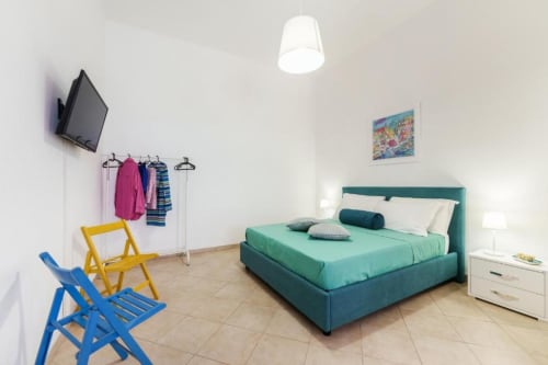 Camera luminosa all'Origenes Rooms di Procida con letto turchese, comodino bianco, TV, sedie colorate e stendibiancheria