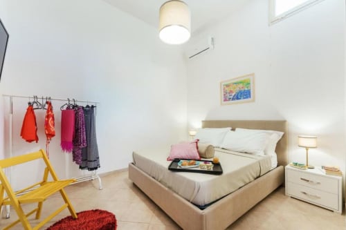 Camera luminosa dell'Origenes Rooms a Procida con letto beige, comodino bianco, sedia gialla e appendiabiti colorato