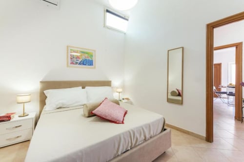 Camera luminosa nell'appartamento di Procida con letto matrimoniale, biancheria bianca, cuscino rosa, cornice in legno, specchio e lucernario