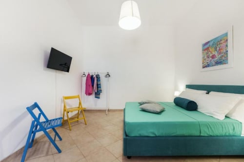 Camera moderna presso Origenes Rooms a Procida con letto verde, sedie colorate, TV e poster con vista sul mare