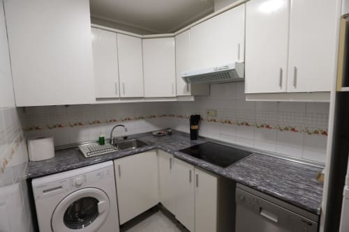 Cocina moderna blanca con encimeras oscuras, estufa, fregadero y lavadora en apartamento de alquiler en Madrid