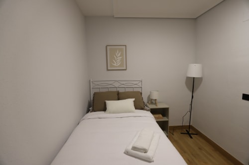 Dormitorio moderno en Innoval Madrid con cama de estructura metálica, ropa de cama blanca, mesita de noche, lámpara de pie y arte botánico en la pared