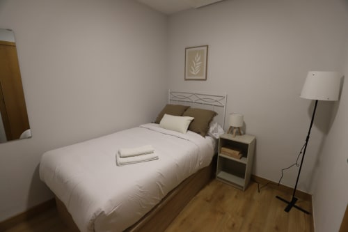 Luminoso dormitorio moderno en Innoval Madrid con ropa de cama blanca, cabecero metálico, mesita de noche de madera, lámpara de pie y detalles botánicos