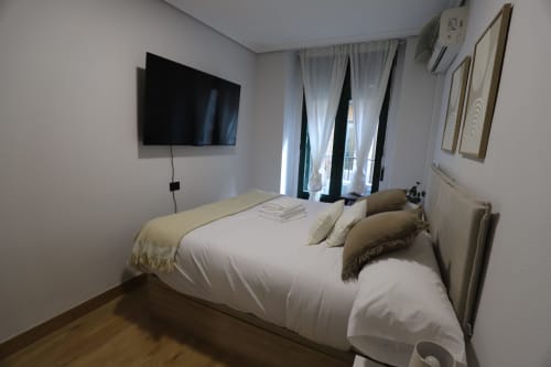 Habitación moderna en Madrid con cama blanca, TV montada en pared y balcón en Innoval Rental Properties