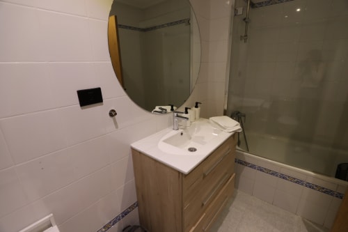 Baño moderno con tocador de madera, espejo redondo, azulejos blancos y cabina de ducha de vidrio en apartamento madrileño