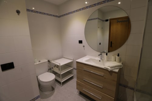 Baño moderno en apartamento madrileño con espejo redondo, tocador de madera, inodoro y paredes de azulejos blancos