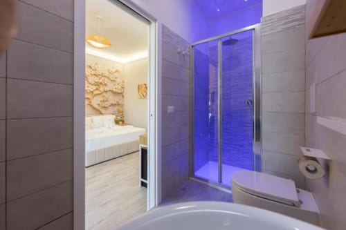 Bagno moderno alle Aurum Nolana Suites di Pompei con box doccia illuminato in blu, wc bianco e vista verso la camera da letto
