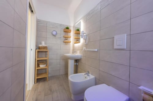 Bagno moderno presso l'Aurum Nolana Suites a Pompei con piastrelle beige, lavabo su colonna, bidet, ripiani in legno e piante in vaso