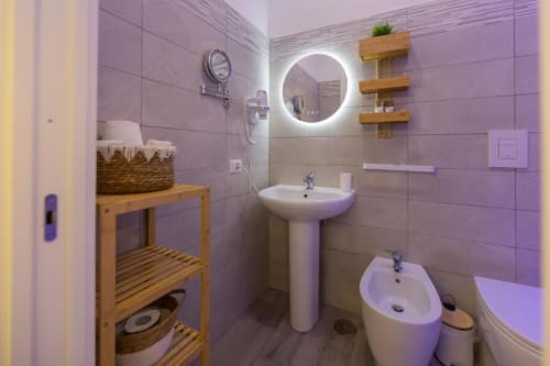 Bagno moderno nelle Aurum Nolana Suites di Pompei con specchio illuminato, lavabo su colonna, bidet, scaffali in legno e accenti viola