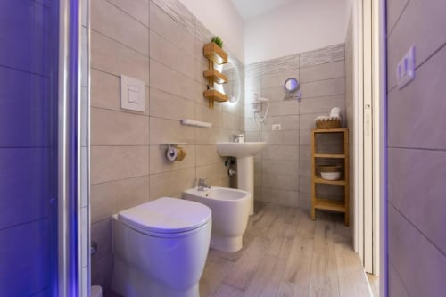 Bagno moderno dell'Aurum Nolana Suites a Pompei con toilette, bidet, lavabo, scaffali in legno e luci ambientali viola