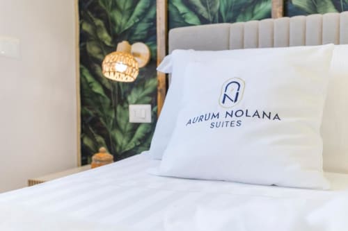 Cuscino bianco con logo Aurum Nolana Suites sul letto, con arte botanica a parete e lampada a sospensione in rattan nella camera dell'hotel Pompeii