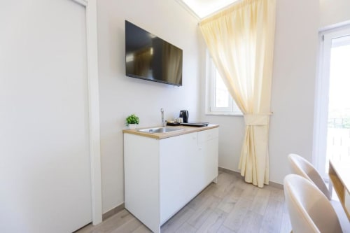 Angolo cottura moderno con mobili bianchi, lavello, TV e tende color panna nella Deluxe Room di Aurum Nolana Suites a Pompei