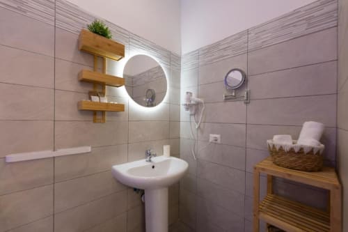 Bagno moderno con specchio illuminato, lavabo bianco su piedistallo, ripiani in legno e piastrelle presso Aurum Nolana Suites a Pompei