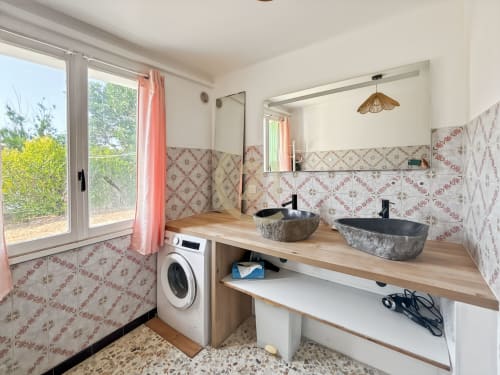 Salle de bain moderne avec vasques en pierre, meuble en bois, lave-linge, carrelage à motifs et vue jardin à Calvi