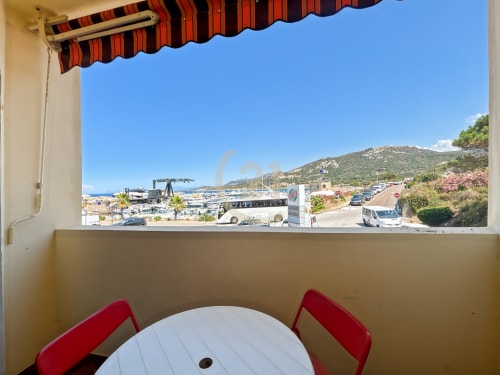 Vue depuis la terrasse d'un appartement avec store rayé rouge et table blanche, donnant sur le port et la plage de L'Île-Rousse