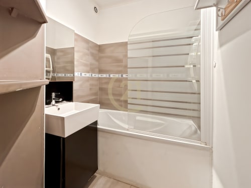 Salle de bain moderne aux carrelages beige, lavabo blanc et baignoire avec paroi vitrée à l'appartement Century 21 de L'Île-Rousse