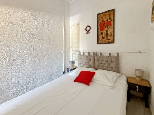 Chambre douillette aux murs blancs, tête de lit capitonnée beige, coussin rouge et tableau vue mer chez Century 21 L'Île-Rousse