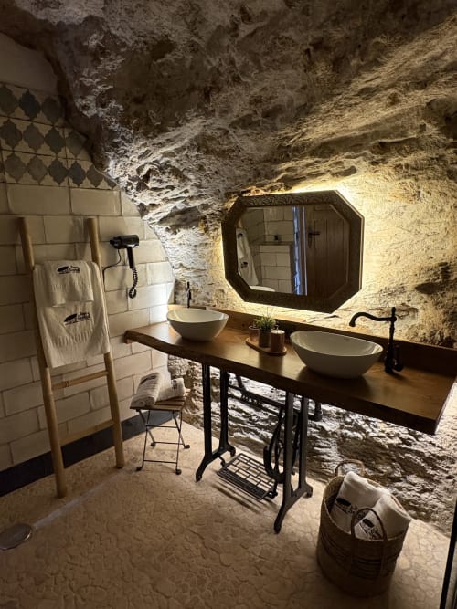 Baño en cueva de La Joya de Setenil con doble lavabo, paredes de piedra iluminadas y decoración rústica