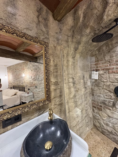 Baño de Casa Cueva Rubí con paredes de piedra, lavabo de mármol negro, espejo ornamentado y ducha de efecto lluvia en La Joya de Setenil