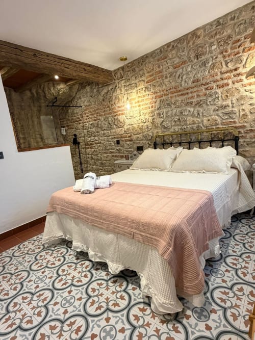 Dormitorio Casa Cueva Rubí con paredes de piedra, cama queen, suelo de azulejos decorativos y vigas rústicas en La Joya de Setenil