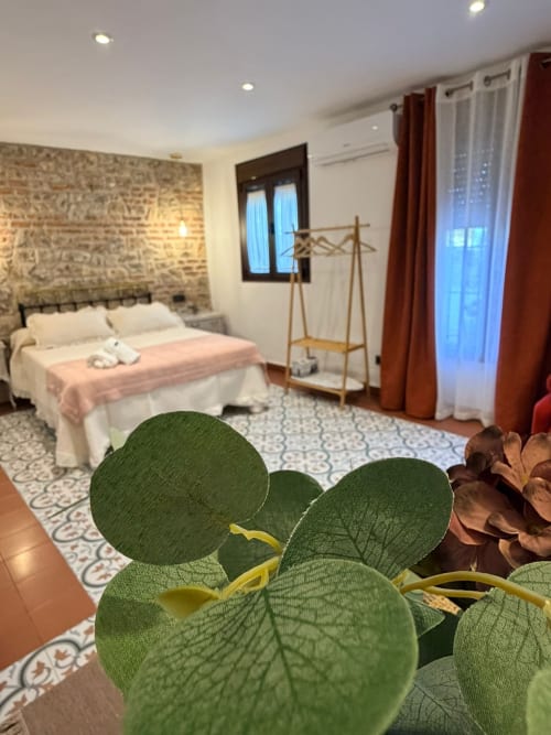 Dormitorio con vistas al jardín en La Joya de Setenil, plantas en macetas, cama con manta rosa y pared de piedra rústica