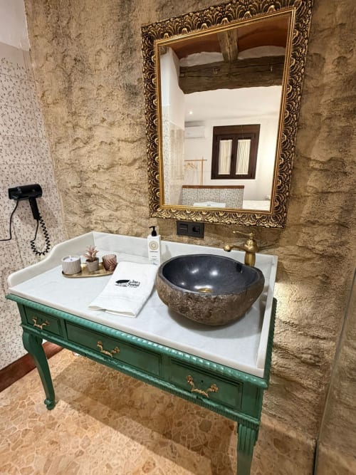 Baño de lujo en apartamento cueva con tocador verde, lavamanos de piedra, espejo ornamental y muros de piedra rústica en La Joya de