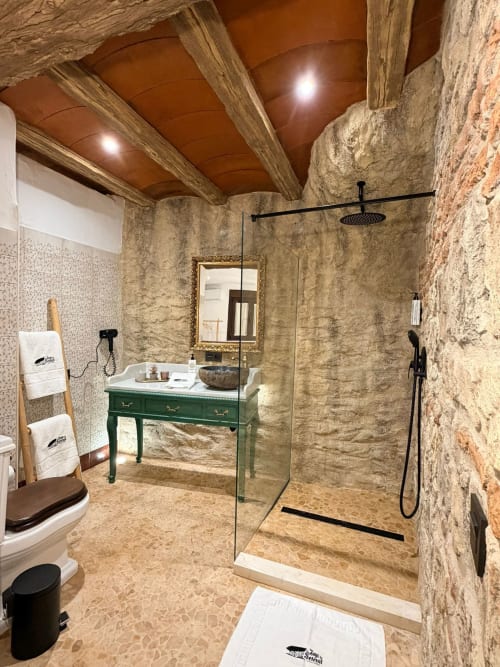 Baño de lujo en cueva de La Joya de Setenil con muros de piedra, tocador verde, ducha de lluvia y techo rústico de madera