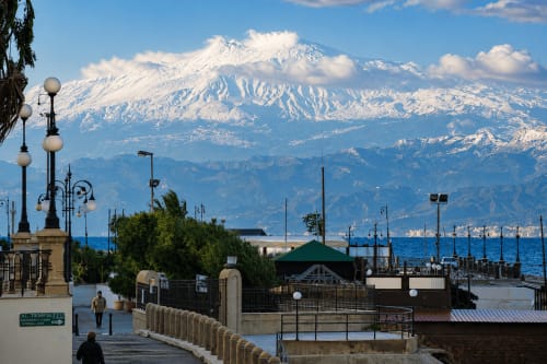Schneebedeckter Ätna vom Uferpromenadeweg in Reggio Calabria mit historischen Laternen und Hafen.