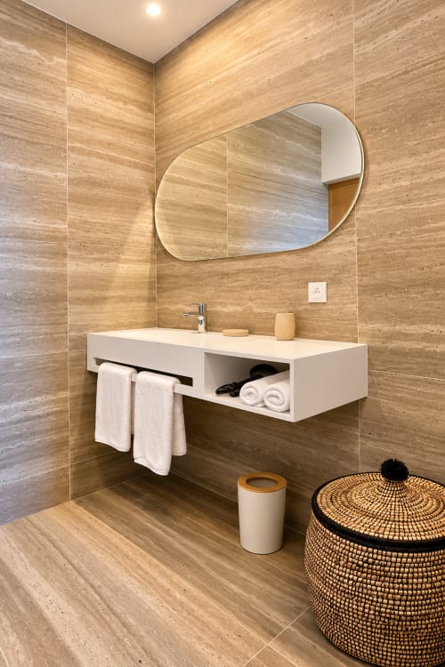 Salle de bain moderne à la Villa Alora avec murs en travertin, vasque flottante blanche, miroir ovale et panier tressé à Calenzana
