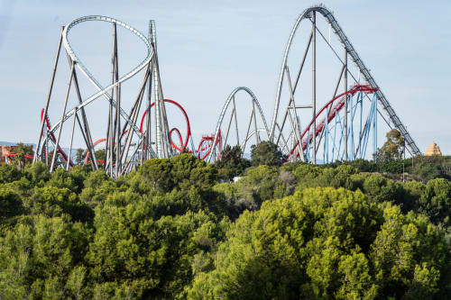 Roller coaster towers with red and blue tracks rising above green trees at Spanish amusement park