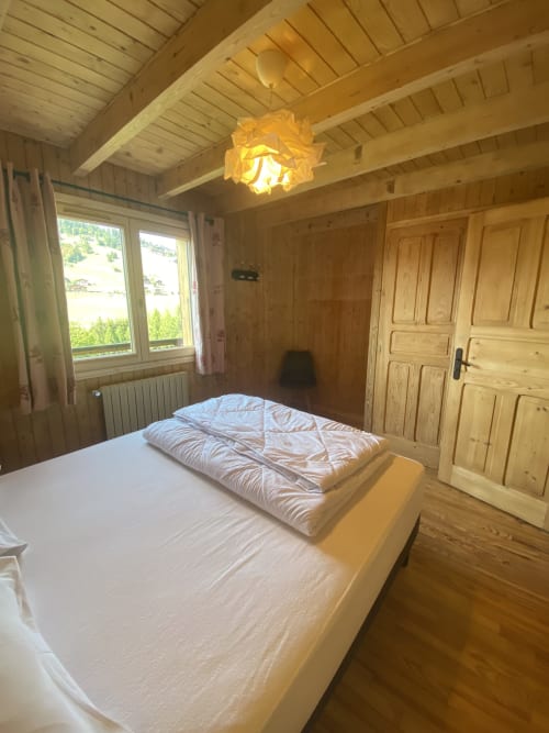Bright wooden bedroom with white bed, yellow pendant light, mountain views through windows, and wooden wardrobe in French