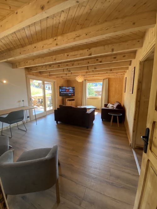 Spacious wooden chalet living room with exposed beams, dark sofas, TV, and mountain views at La Ferme des Georgières