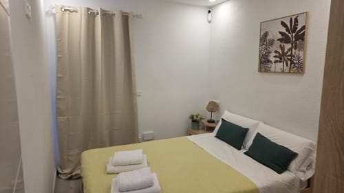 Dormitorio luminoso en la casa rural de Benicàssim con colcha amarilla, almohadas blancas, cojines verdes, cortinas crema y pared tropical
