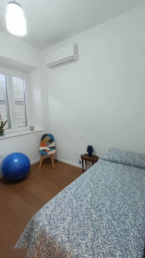 Camera moderna a Roma Casa Tua con letto, palla fitness blu, sedia colorata, pavimenti in legno, climatizzatore e ampia finestra