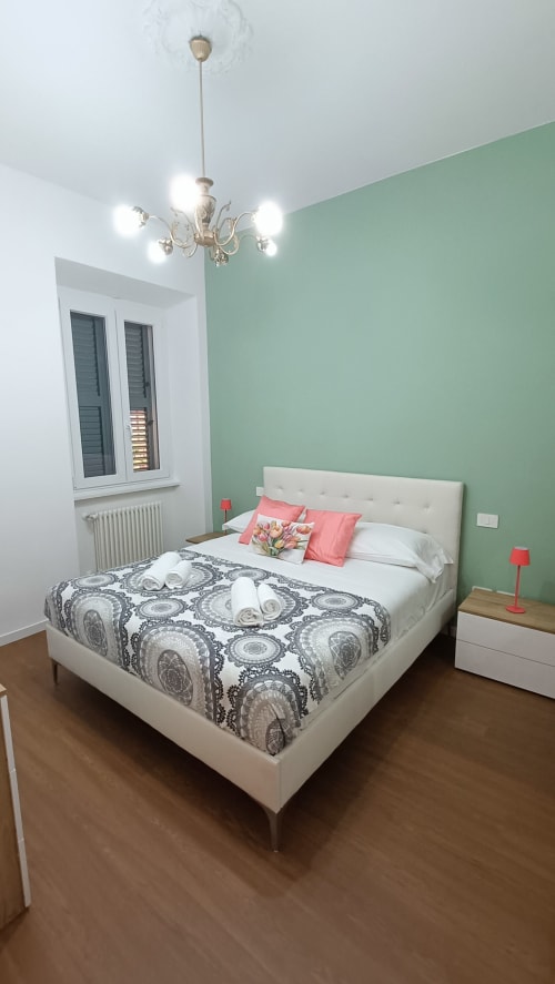Camera luminosa di Roma Casa Tua con parete accent verde menta, letto bianco, lampadario ornamentale e pavimento in legno