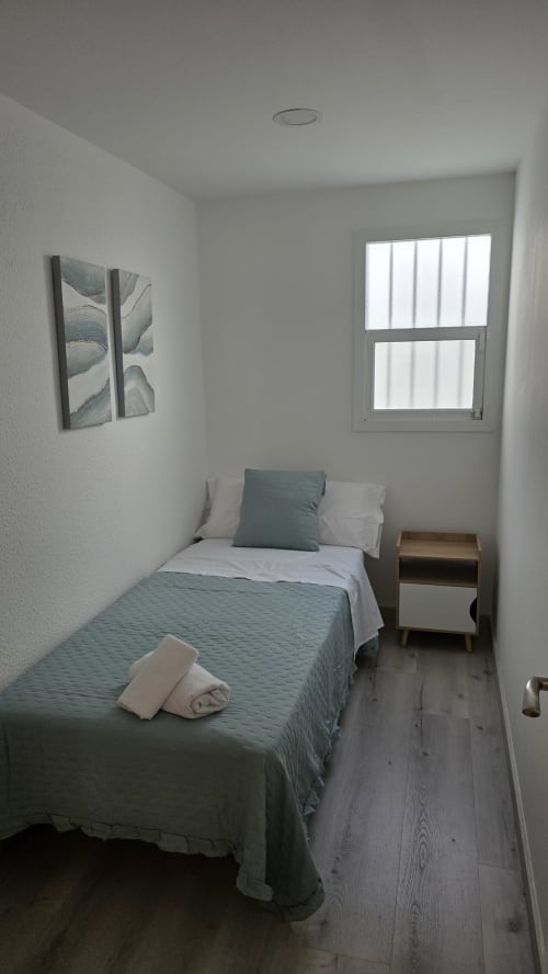 Dormitorio minimalista en casa rural de Benicàssim con cama individual, ropa de cama gris, mesita de madera, ventana y arte botánico