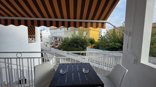 Balcón de apartamento en Benicàssim con toldo rayado, mesa negra de comedor, vistas al jardín y edificios coloridos cercanos