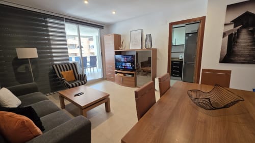 Sala de estar moderna del apartamento frente al mar en Benicàssim con sofá, televisión, mesa de comedor de madera y balcón con vistas al mar