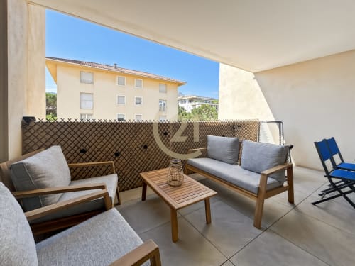 Terrasse ensoleillée à Calvi avec mobilier en bois, coussins gris, vue sur la ville et architecture méditerranéenne