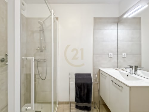 Salle de bain moderne avec douche à l'italienne, vasque blanche et serviette noire dans un appartement Century 21 à Calvi, France