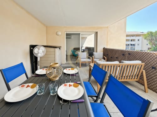 Terrasse ensoleillée de l'appartement à Calvi avec table de salle à manger, chaises bleues, coin salon et vue sur la ville