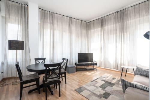 Soggiorno moderno a Milano con tavolo da pranzo nero, TV, tende trasparenti, pavimenti in legno e arredi contemporanei