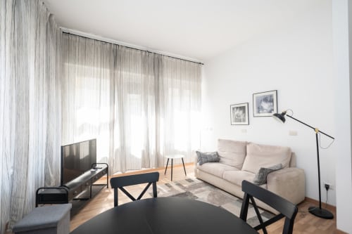 Soggiorno di un moderno appartamento a Milano con divano beige, televisore, arredi neri e tende trasparenti che lasciano filtrare la luce naturale